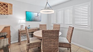 A dining area with wicker chairs, a round table, a hanging light fixture, a television, and a window with blinds. - Blue Surf 5 (Miramar Beach)