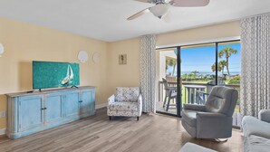 A spacious living room with a wooden floor, a ceiling fan, a blue cabinet, a patterned armchair, a leather armchair, and a large window offering a view of the ocean.