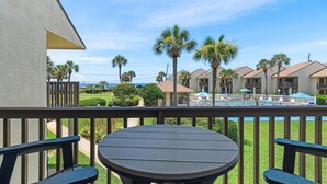 A wooden deck with a round table and benches, overlooking a pool area with palm trees and a view of a resort building. - Blue Surf 7 (Miramar Beach)
