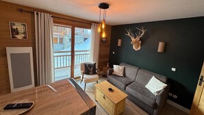 Ein gemütliches Wohnzimmer mit einem Holztisch, einem grauen Sofa und einem Couchtisch aus Holz. Ein großes Fenster mit Vorhängen, ein aufgehängter Hirschkopf an der Wand und eine Deckenleuchte sind ebenfalls vorhanden.