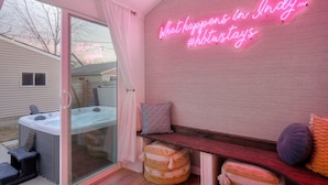 A cozy room with a glass door leading outside, a neon sign on the wall, a hot tub, and a bench with cushions.