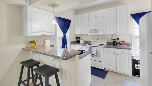 A kitchen with white cabinets, a granite countertop, a microwave, an oven, and a sink. There are two black bar stools and a small island with a plant. - Minutes from Cherry Point (Havelock)