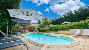 A swimming pool with lounge chairs and umbrellas, surrounded by greenery and a wooden fence.