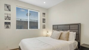 A bedroom with a bed, a nightstand, a wall-mounted lamp, and a window with blinds.