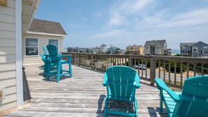 Una terraza de madera con sillas Adirondack turquesa, barandilla de madera y vistas a casas de playa.