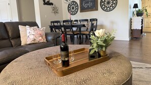 Una sala de estar con un sofá de cuero marrón oscuro, una mesa redonda de centro con una botella de vino y copas, una zona de comedor con una mesa y sillas de madera, y decoración en la pared.