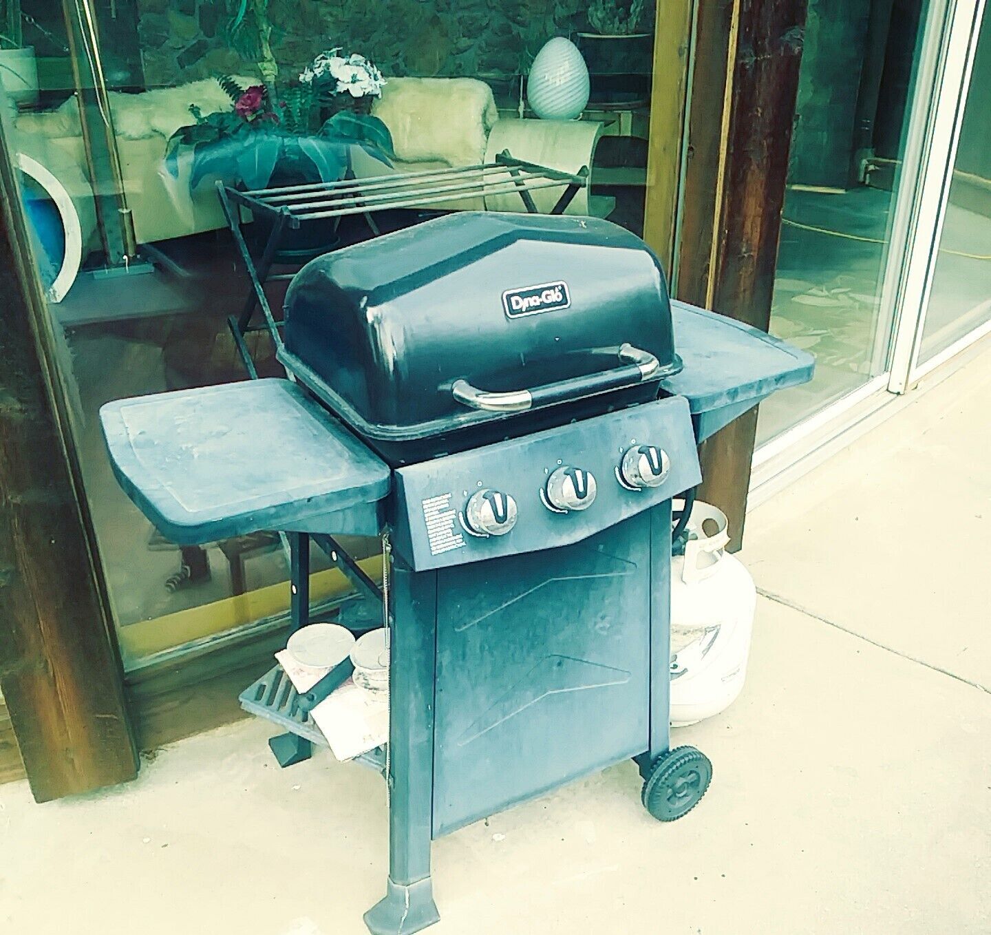 A portable grill with four burners and a side table, placed outside a building with a glass door.