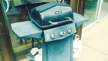 A portable grill with four burners and a side table, placed outside a building with a glass door.