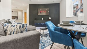 Una sala de estar moderna con un sofá gris, sillas azules y una mesa de comedor preparada para cuatro. Un televisor de pantalla plana montado en una pared oscura muestra el logotipo de Netflix.