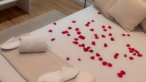 Cama bem arrumada com pétalas de rosa, um guarda-roupa branco e uma pequena mesa de cabeceira.