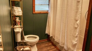Ein Badezimmer mit Holzboden, Toilette, Ablagefläche mit Handtüchern, Fenster und Duschvorhang.