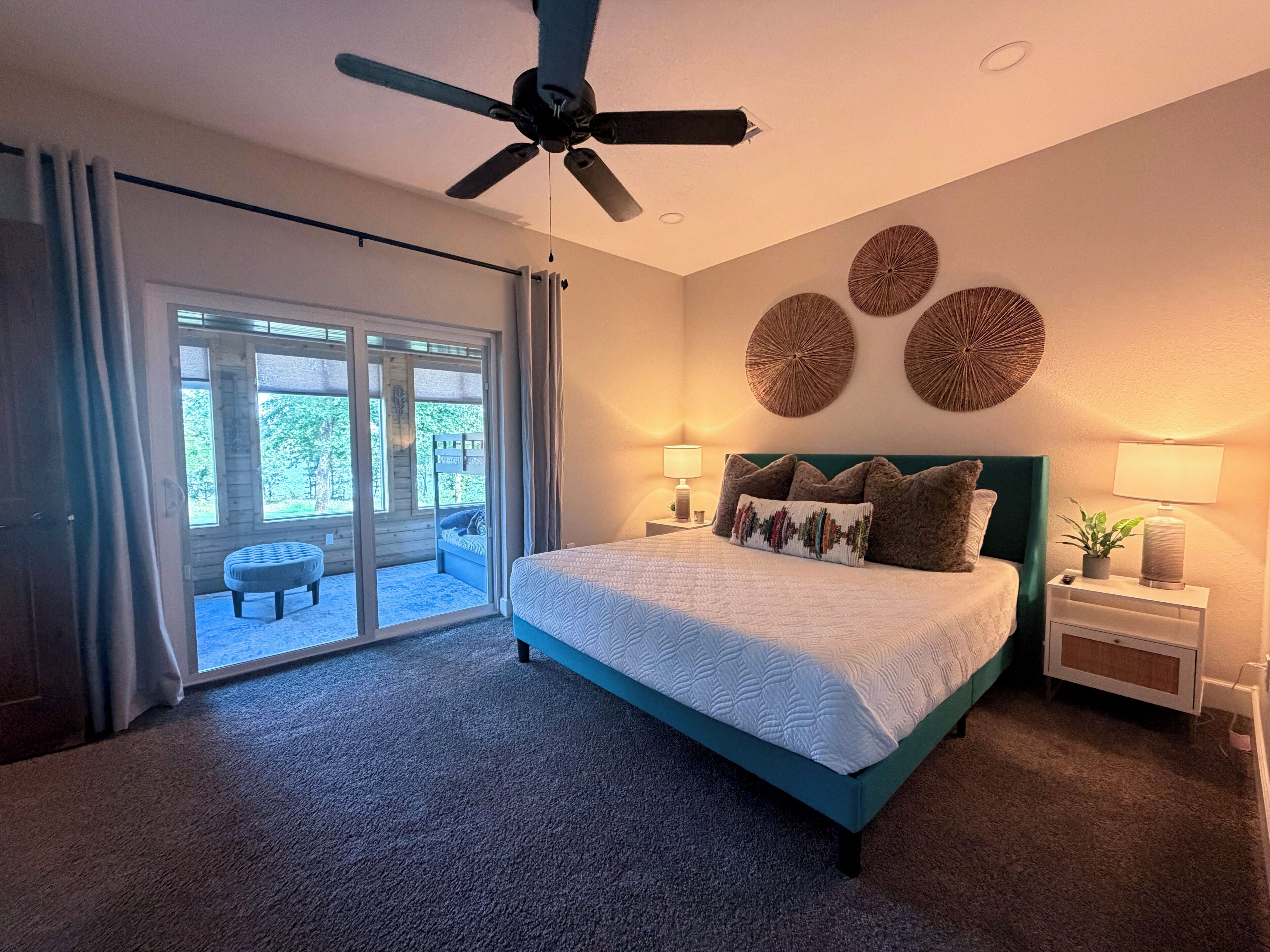 Une chambre à coucher avec un lit, un ventilateur de plafond, une table de chevet et une vue sur l’extérieur, accessible par une porte-fenêtre coulissante.