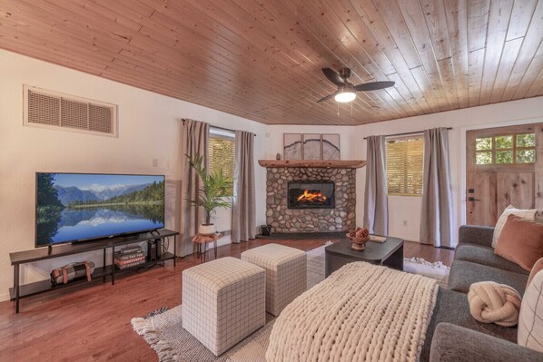 A cozy living room with a stone fireplace, a flat-screen TV, wooden flooring, and a ceiling fan.