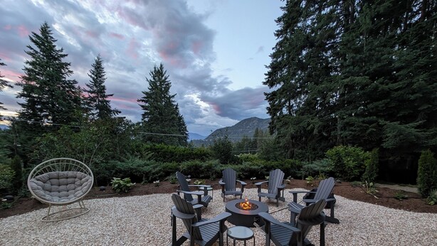 An outdoor seating area with Adirondack chairs and a round table with a fire pit, surrounded by gravel and dense trees.