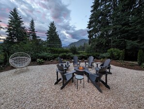 Un espacio al aire libre con sillas Adirondack y una mesa redonda con un brasero, rodeado de grava y árboles frondosos.
