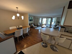 A modern kitchen with a dining area, wooden flooring, and a large window providing natural light.