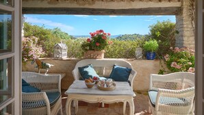 Un patio cubierto con mobiliario de mimbre, una mesa con una bandeja de pan y fruta, y vistas a zonas verdes y colinas.