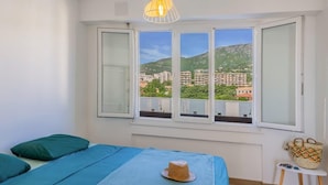 Un dormitorio con cama, ventana con vistas a edificios y una colina, y una lámpara colgante.