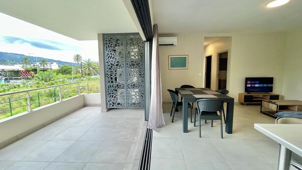 A balcony with a view of greenery and buildings, a dining area with chairs and a table, and a TV on a stand.