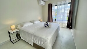 A hotel room with a bed, a bedside lamp, an air conditioning unit, a window with curtains, and a view of buildings outside.