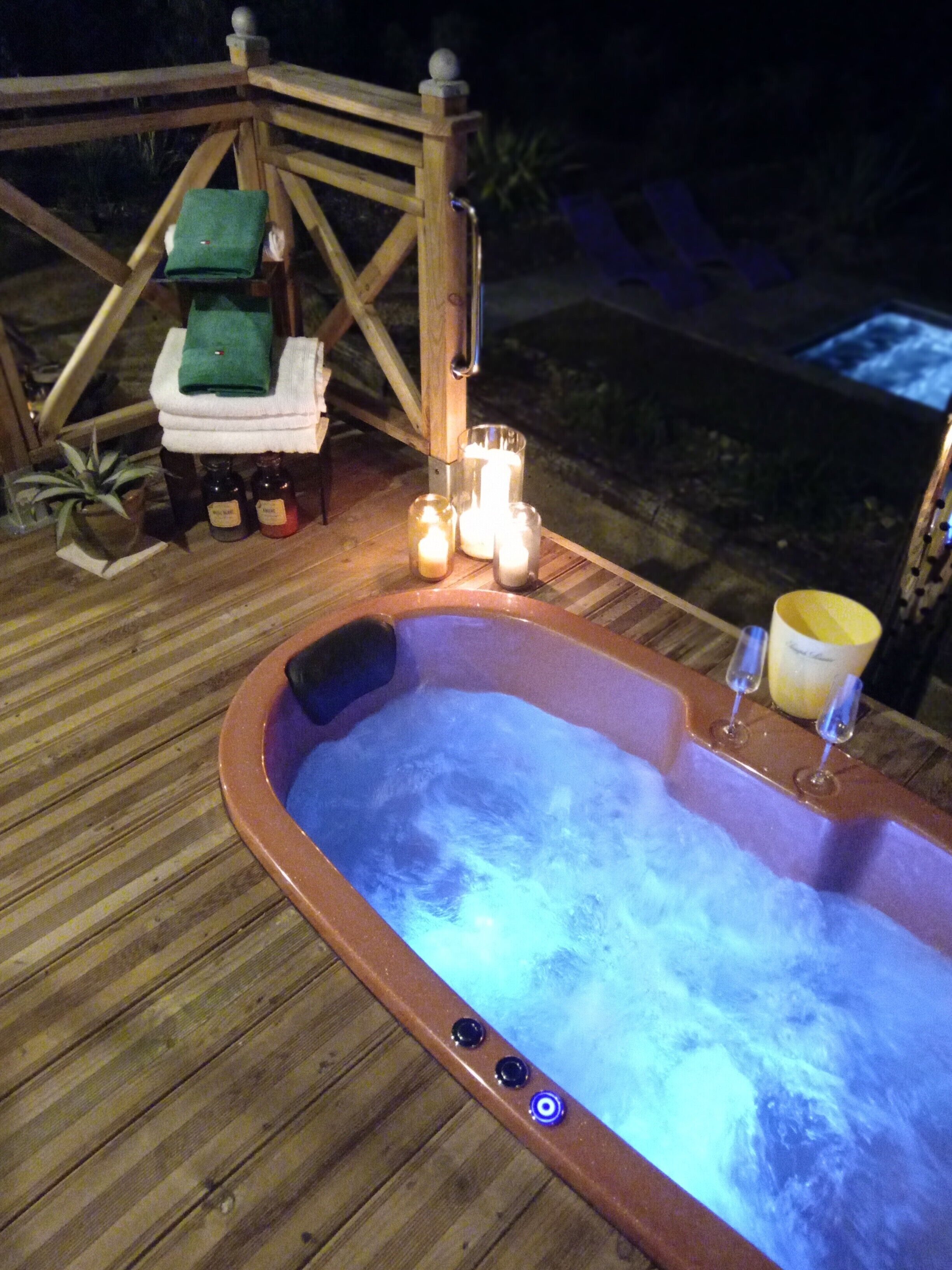 Un jacuzzi con velas y botellas, una terraza de madera y una pequeña mesa con toallas.