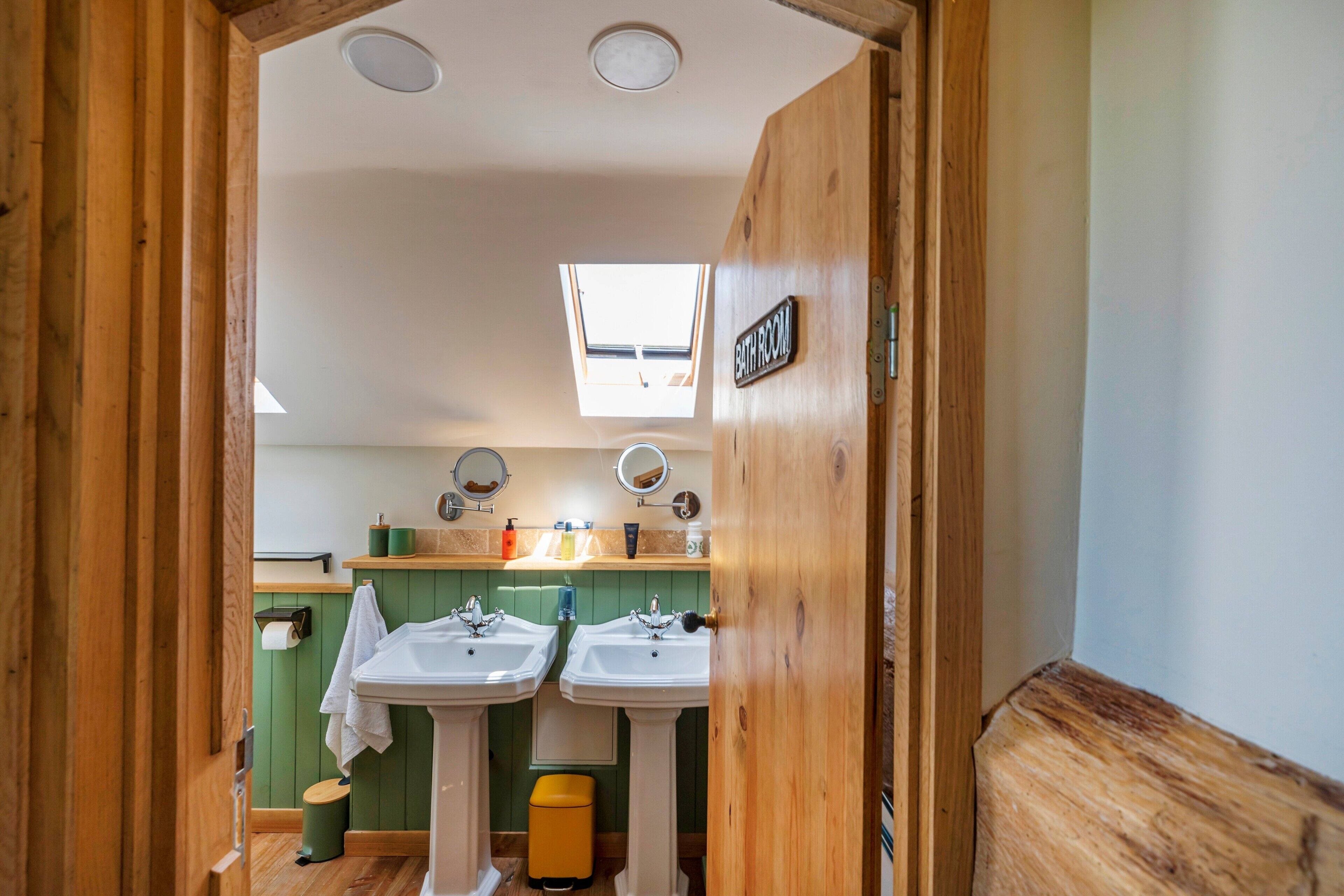 A bathroom with two sinks, a toilet, a trash bin, and a skylight.