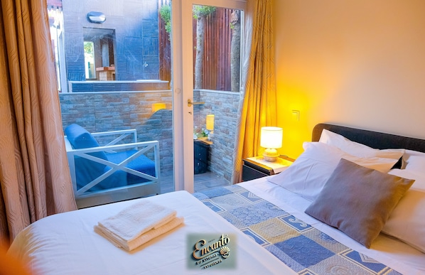 Une chambre d’hôtel comprenant un lit, une chaise, une table de chevet avec une lampe, et donnant sur un espace extérieur agrémenté d’un mur en pierre et d’une clôture en bois.