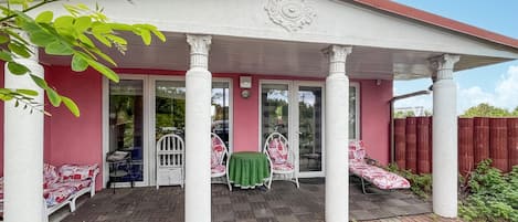 A covered patio with two white chairs and a table, a red fence, and a brick pathway.