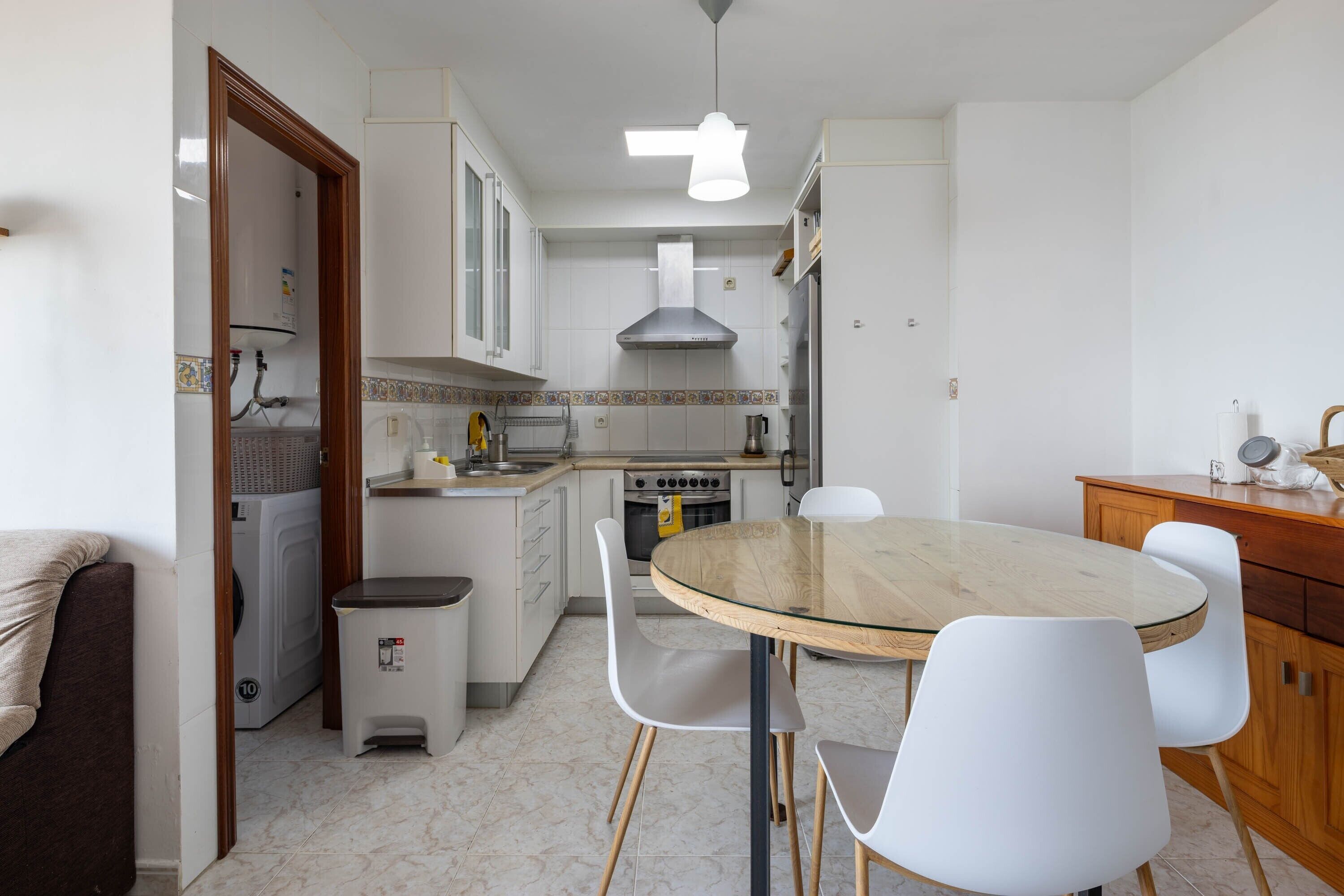 Una cocina funcional con gabinetes blancos, estufa, lavamanos y comedor con mesa de vidrio y sillas blancas.