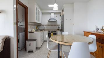 Una cocina funcional con gabinetes blancos, estufa, lavamanos y comedor con mesa de vidrio y sillas blancas.
