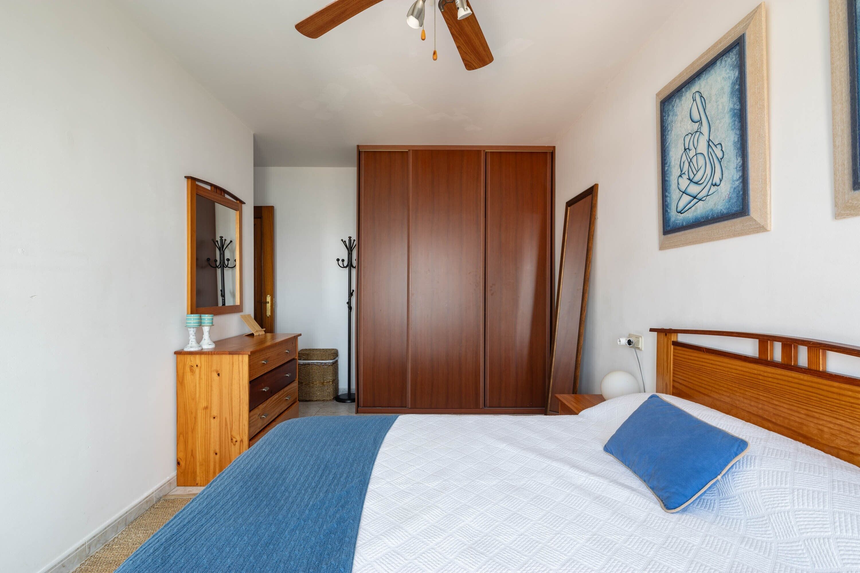 Quarto com cama, guarda-roupa de madeira, ventoinha no teto, obra de arte na parede e mesa de cabeceira.