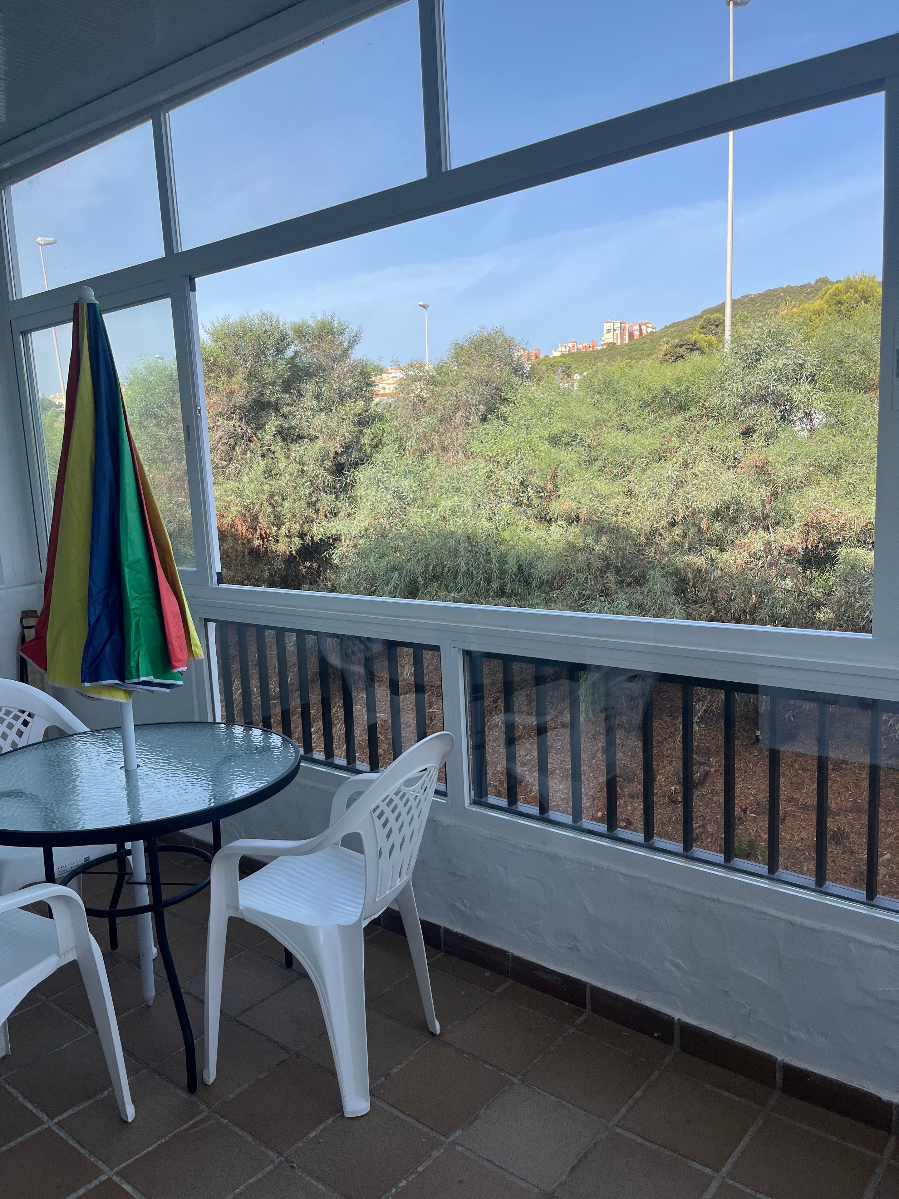 A glass-enclosed balcony with a table and chairs, a colorful umbrella, and a view of a hilly landscape.