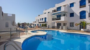 A swimming pool area with a clear blue pool, white buildings with blue accents, and a sunshade structure.