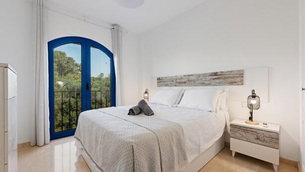 A bedroom with a bed, a nightstand, a lamp, a window with curtains, and a view of greenery outside.