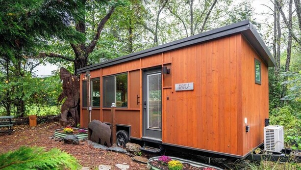 Una cabaña rústica de madera con un letrero que dice "Rusty's Retreat". Cuenta con ventanas grandes, una puerta y está rodeada de un jardín con plantas y un árbol grande.