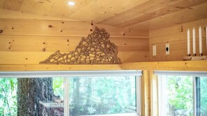 Interior de una cabaña con paredes de madera, chimenea de piedra y ventanas con vistas a un bosque.
