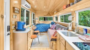 Una cabaña de madera compacta y bien iluminada con kitchenette, una cama con colcha estampada azul y una ventana con vistas a la vegetación.