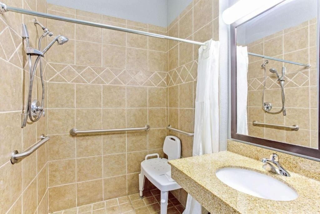 Une salle de bain carrelée de carreaux beiges, une douche équipée d’une barre d’appui, des toilettes blanches et un lavabo avec un miroir.