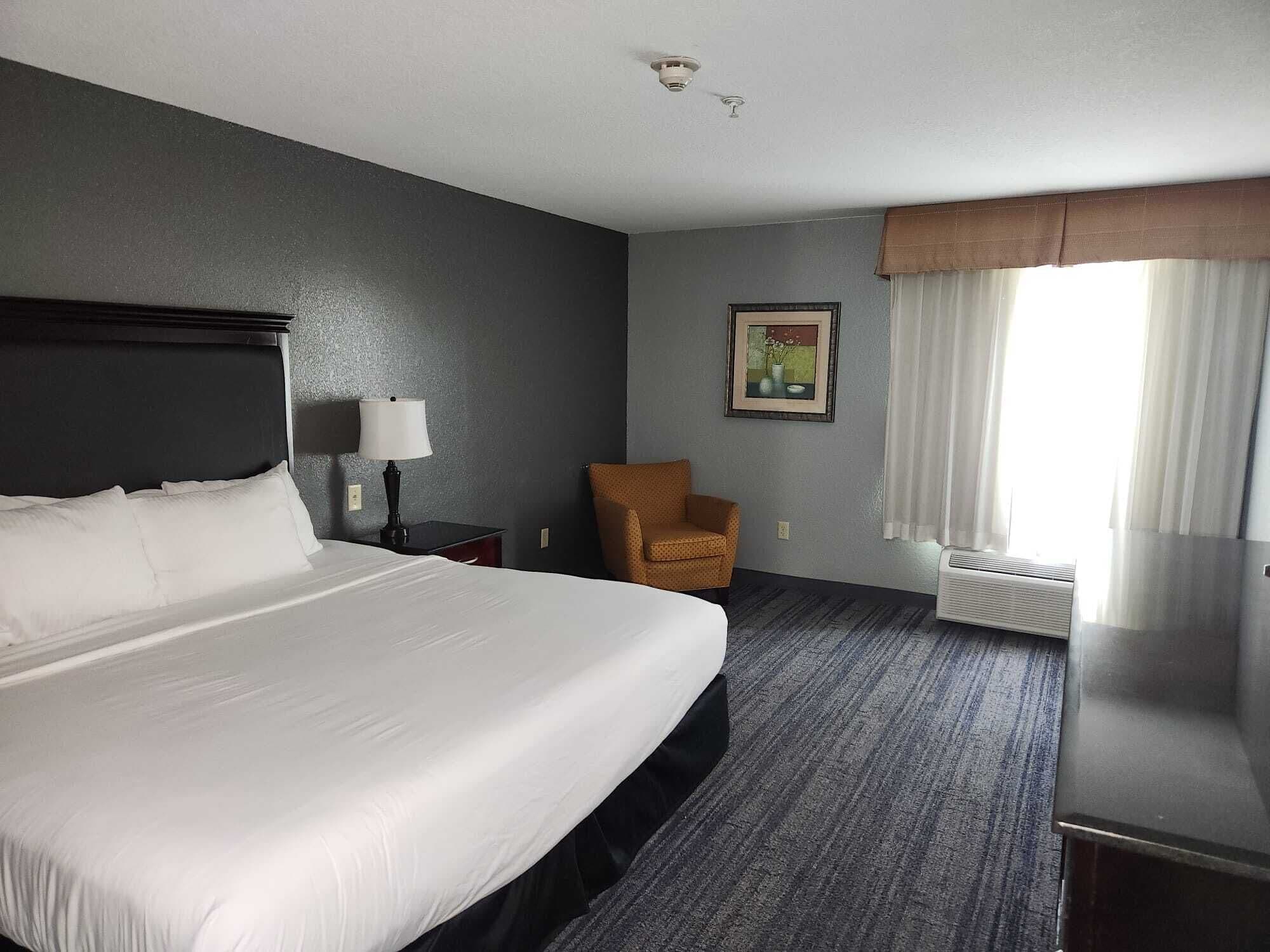 A hotel room with a large bed, a chair, a nightstand, a lamp, a painting, and a window with curtains.