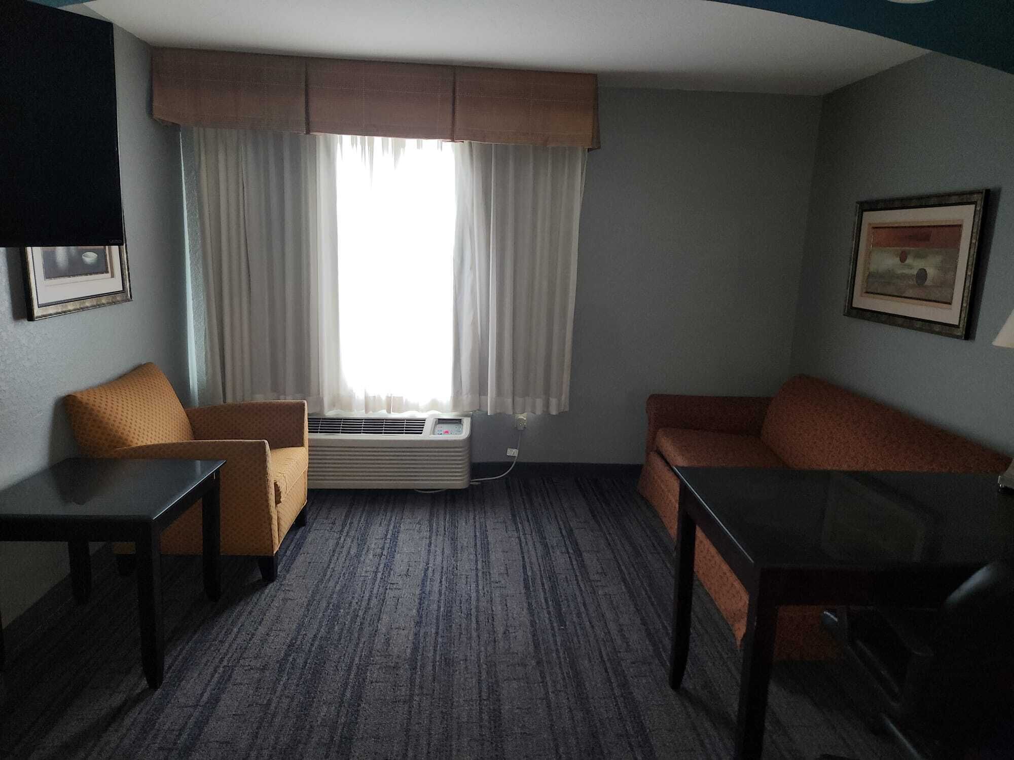Une petite chambre d’hôtel, de taille modeste, comprenant un canapé, une chaise, une petite table et une fenêtre avec des rideaux.