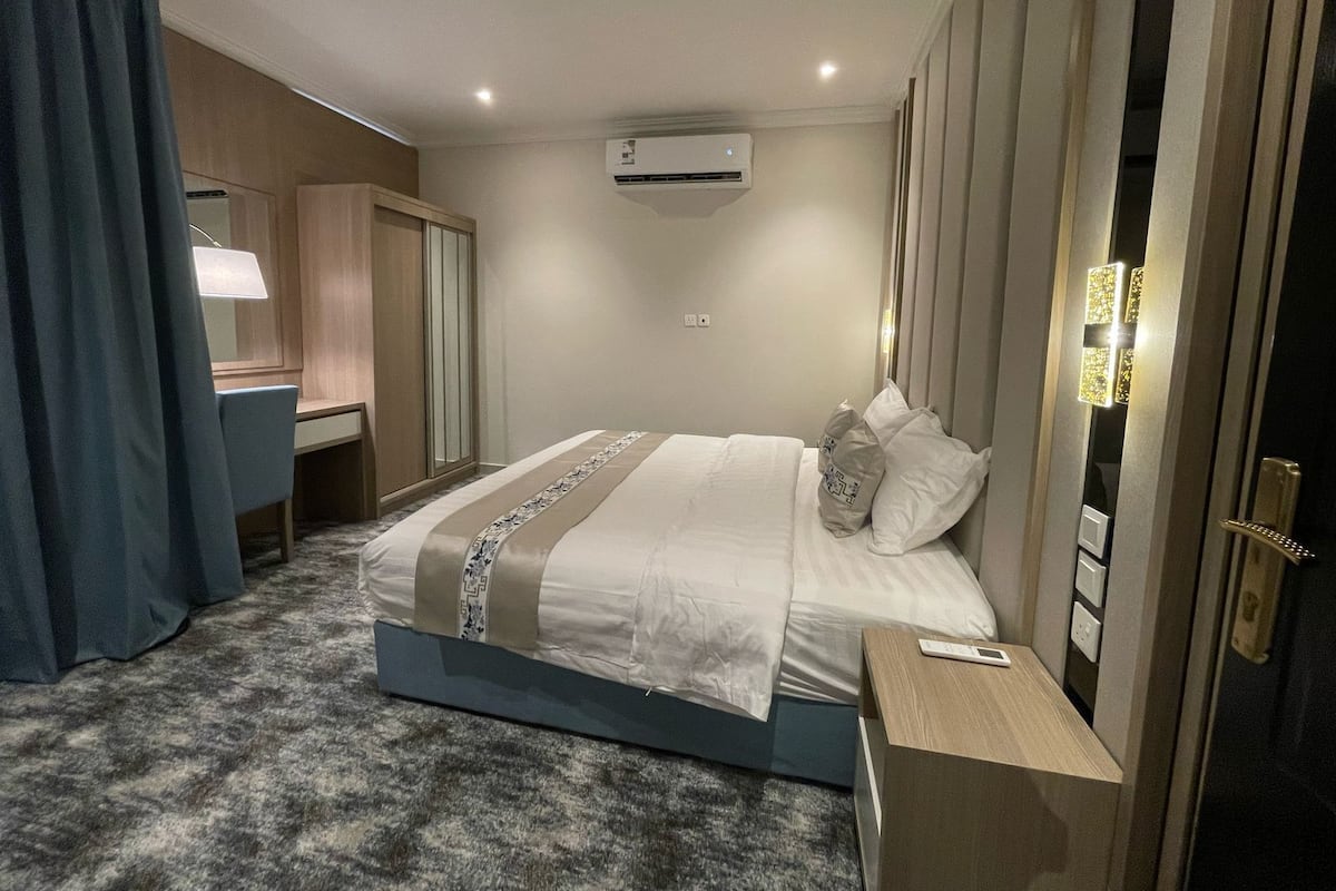 A hotel room with a bed, bedside table, lamp, wardrobe, and an air conditioning unit.