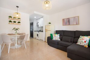Uma sala moderna com área de refeições, um sofá com almofadas decorativas, cozinha ao fundo e luminária pendente.