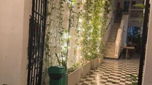 Een smalle gang met een zwart-wit geruit vloerkleed, een metalen poort aan de linkerkant, en een muur versierd met klimplanten en lichtjes.