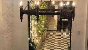 Een smalle gang met een zwart-wit geruite vloer, een decoratieve boog met hangende lampen en potplanten aan weerszijden.