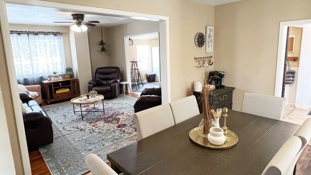 A dining area with a dark wooden table, white chairs, and a decorative centerpiece. There is a living room in the background with a sofa, coffee table, and a ceiling fan. - Sturgis Rally perfect location! (Sturgis)