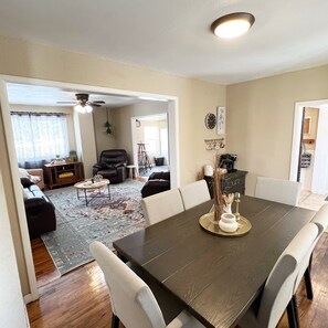 A dining area with a dark wooden table, white chairs, and a decorative centerpiece. There is a living room in the background with a sofa, coffee table, and a ceiling fan. - Sturgis Rally perfect location! (Sturgis)