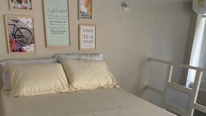 Ein ordentlich bezogenes Bett mit einer beigen Bettdecke und Kissen, eine Wand mit gerahmten Kunstwerken und ein Fenster mit weißem Rahmen.