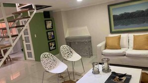 Ein modernes Wohnzimmer mit einem weißen Sofa, zwei weißen Sesseln, einem Couchtisch und einem Kamin. Im Hintergrund ist eine Treppe mit weißer Geländer zu sehen.