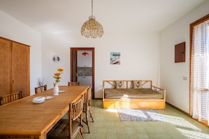 Una habitación con una mesa de comedor de madera, sofá cama, lámpara colgante, zona de cocina y ventana con cortina.
