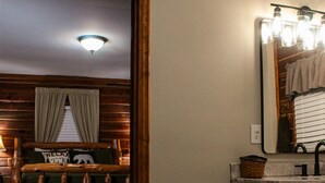 A room with a wooden bed, a bathroom with a mirror and lights, and a small basket by the door.